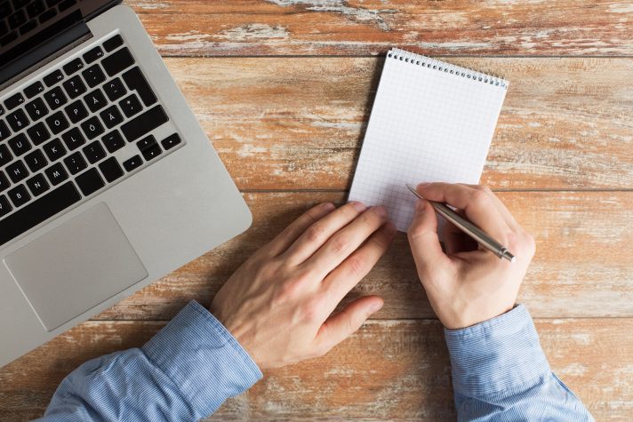 Image of Caucasian male taking notes on a notepad with a laptop beside him