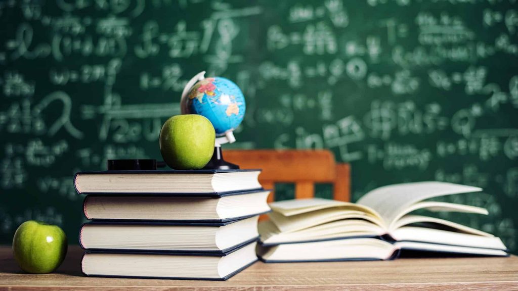 textbooks and other educational material on top of a desk in a classroom with a chalkboard behind it