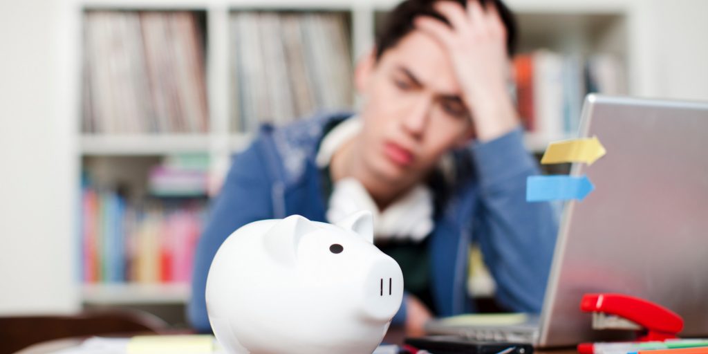 a stressed male student looking despairingly at a piggy bank on his desk